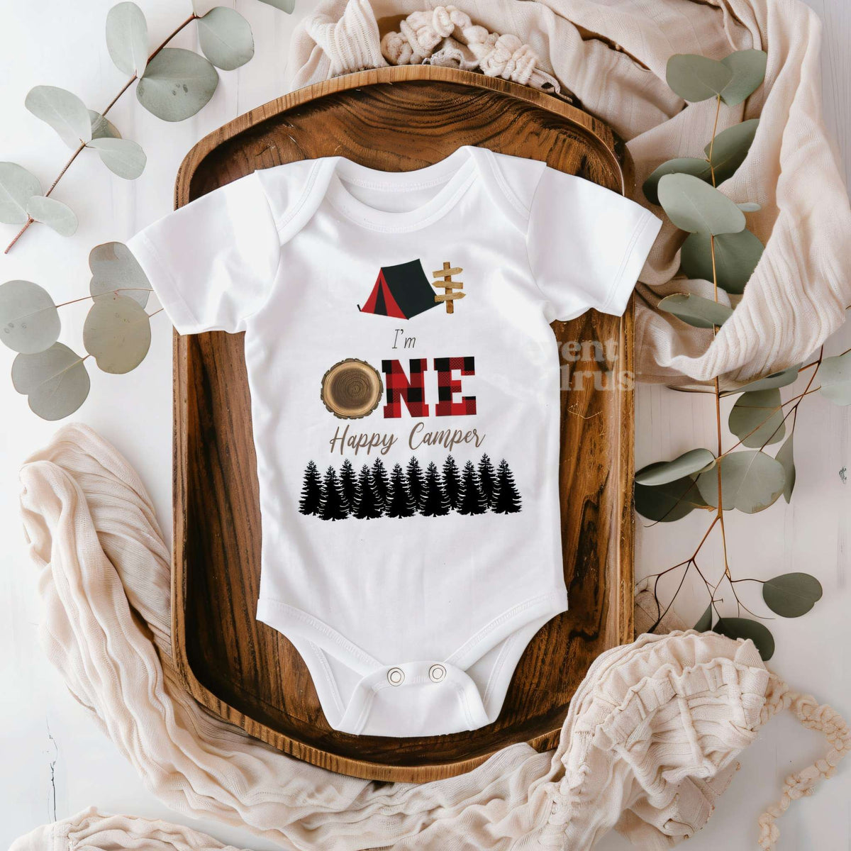 White I'm One Happy Camper bodysuit on a wooden tray with eucalyptus and a knit blanket.