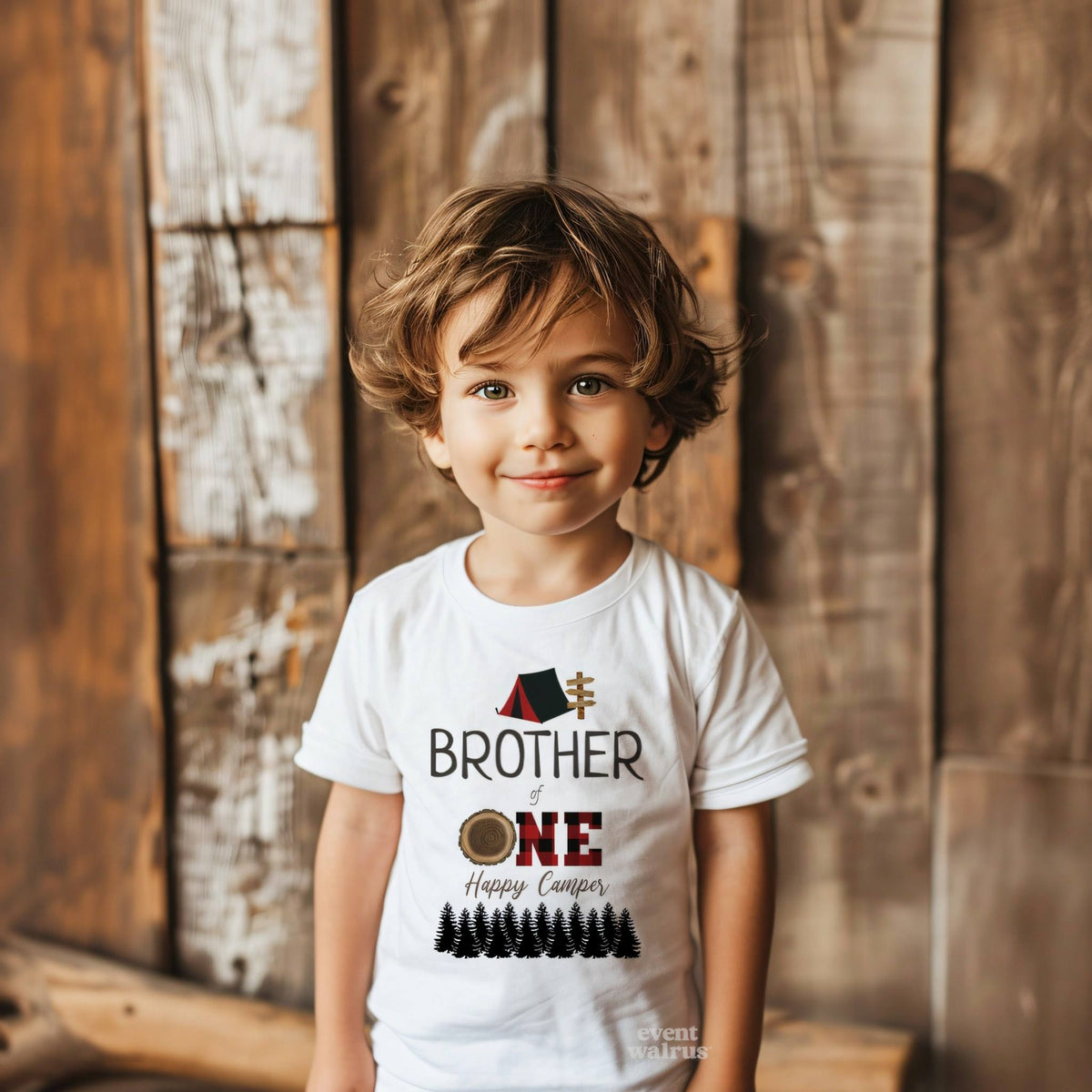 Toddler boy against a weathered wood wall in a white Brother of One Happy Camper tee.