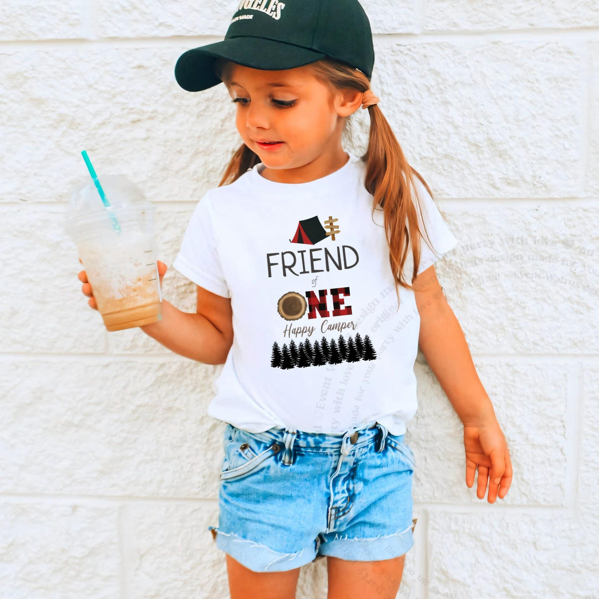 Girl in a ball cap holding a drink in a white Friend of One Happy Camper shirt.
