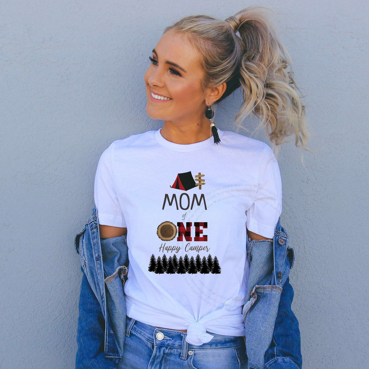Smiling woman in a denim jacket wearing a white red plaid Mom of One Happy Camper shirt.