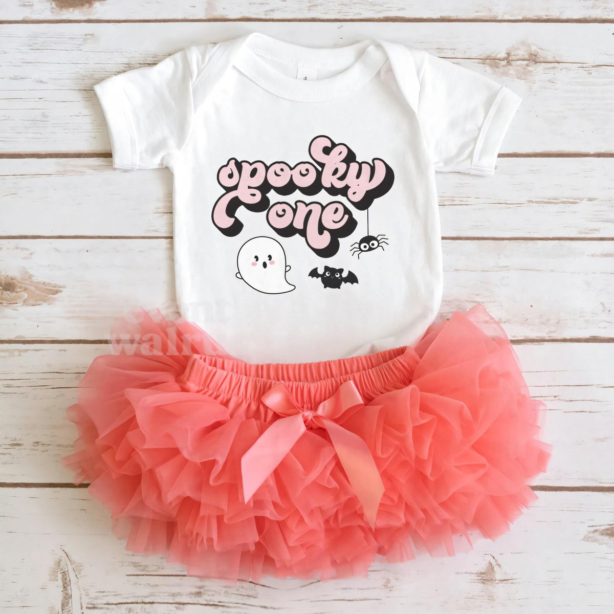 White Spooky One bodysuit laid flat with a coral tutu on whitewashed wood.