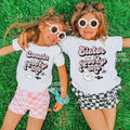 Two girls in heart sunglasses lying on grass in white Cousin and Sister Spooky One shirts.