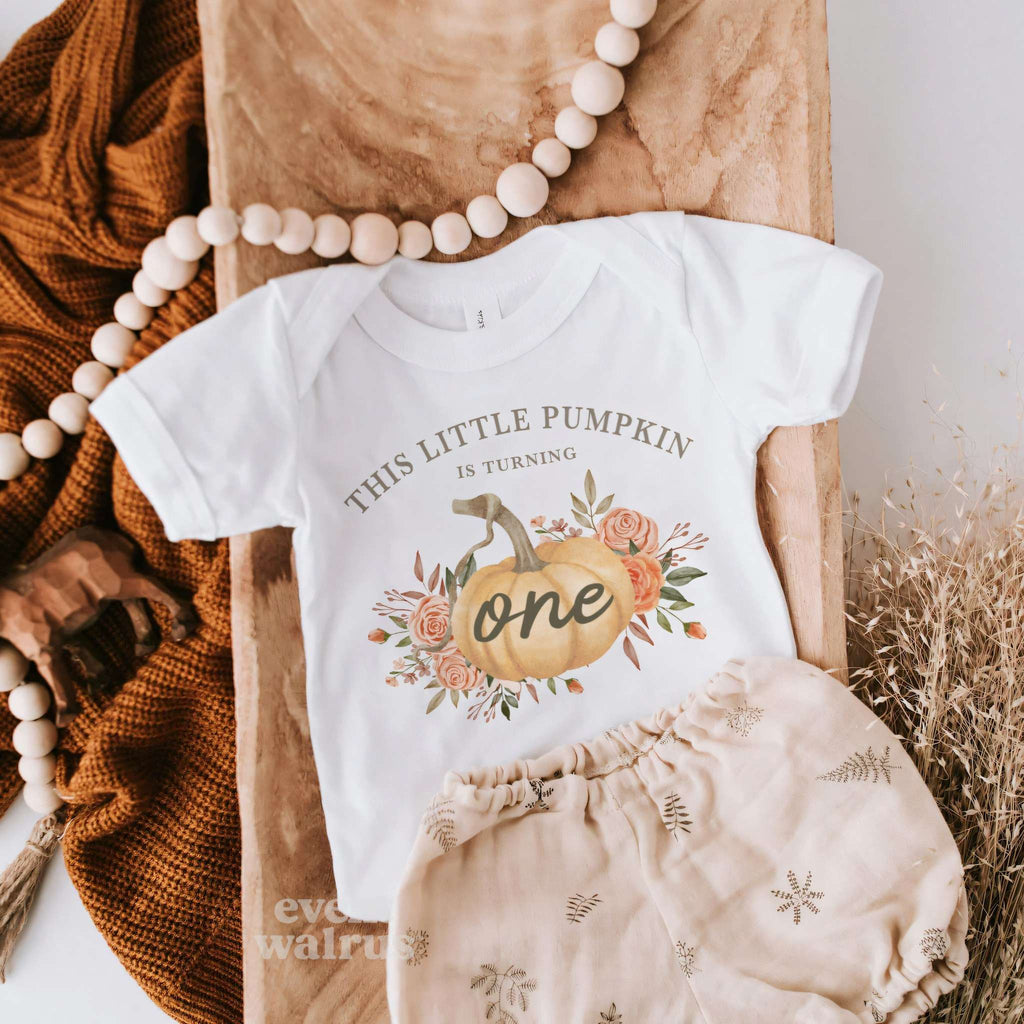 White rustic Little Pumpkin bodysuit laid flat with a knit blanket and dried grasses.
