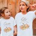 Two girls against a wood wall in white Cousin and Sister rustic Little Pumpkin shirts.