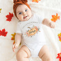 Baby girl in a rust headband lying among fall leaves in a white This Little Pumpkin Is Turning One bodysuit.