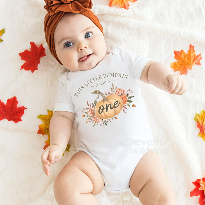 Baby girl in a rust headband lying among fall leaves in a white This Little Pumpkin Is Turning One bodysuit.