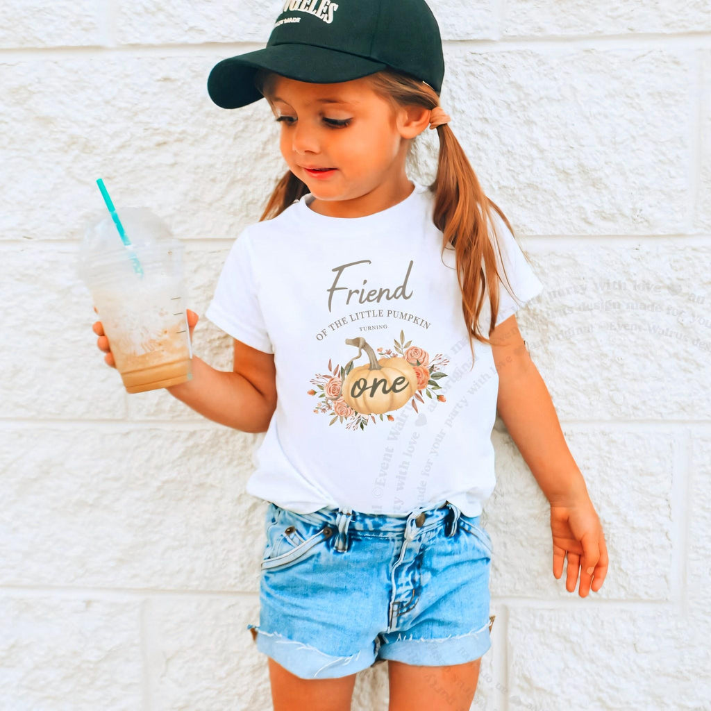 Girl in a ball cap holding a drink in a white Friend of the Little Pumpkin shirt.