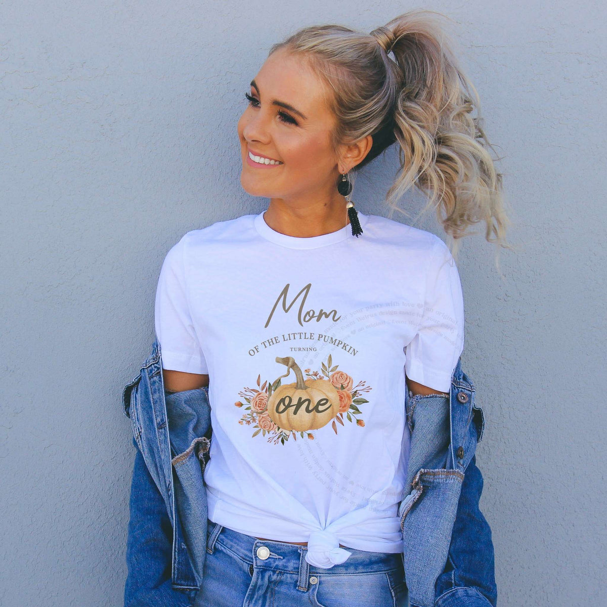 Smiling woman in a denim jacket wearing a white Mom of the Little Pumpkin rustic shirt.