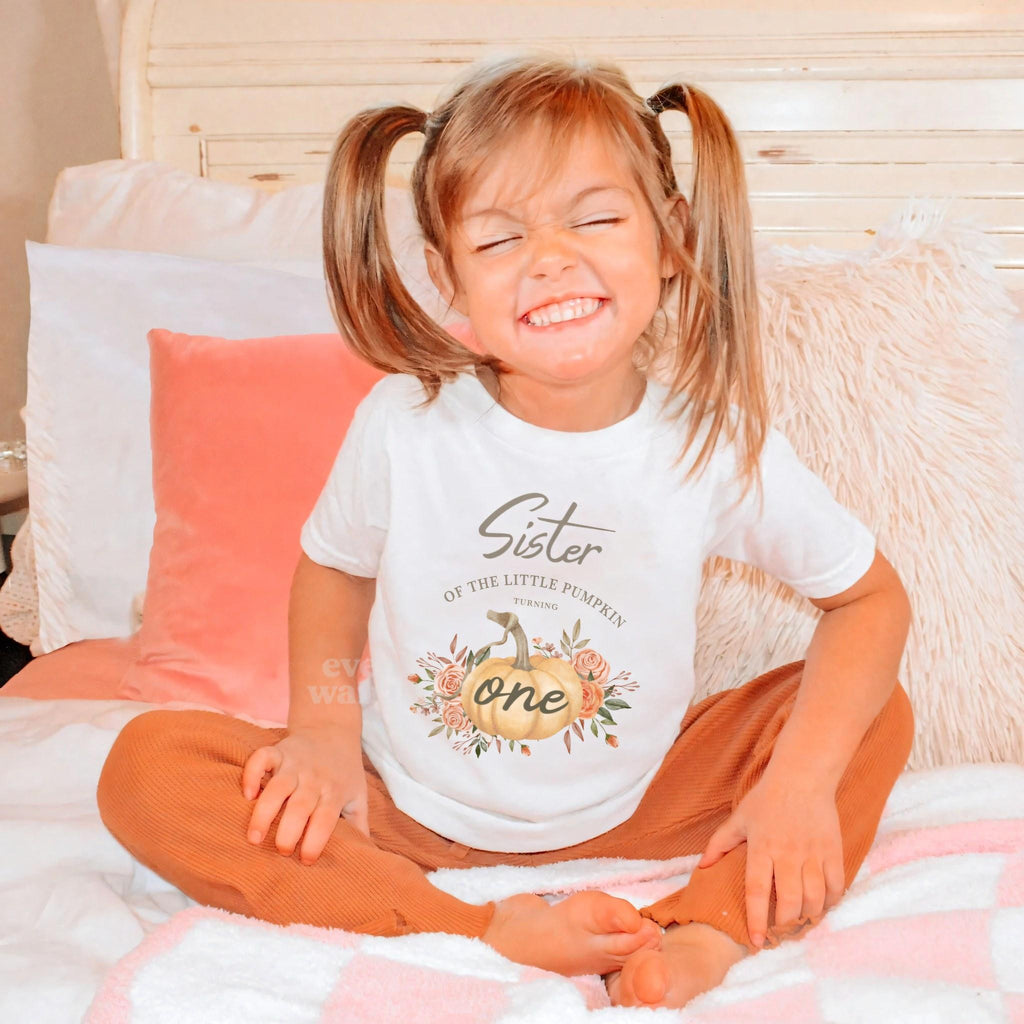 Toddler girl with pigtails sitting in a white Sister of the Little Pumpkin tee.