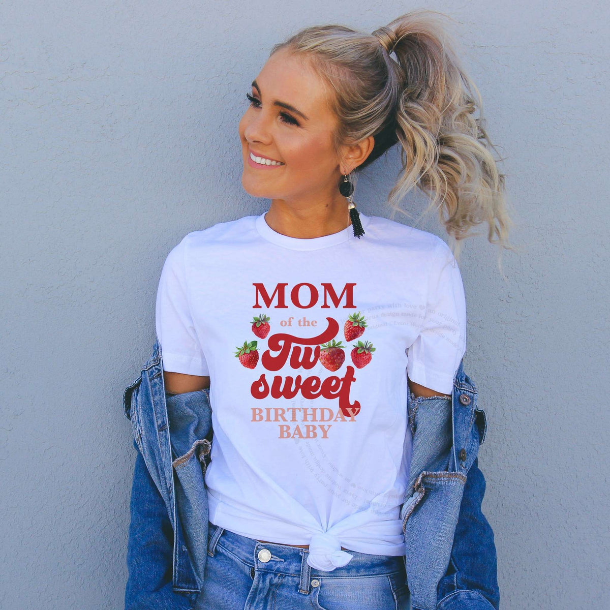 Smiling woman in a denim jacket wearing a white Mom of the Two Sweet shirt with red watercolor strawberries.