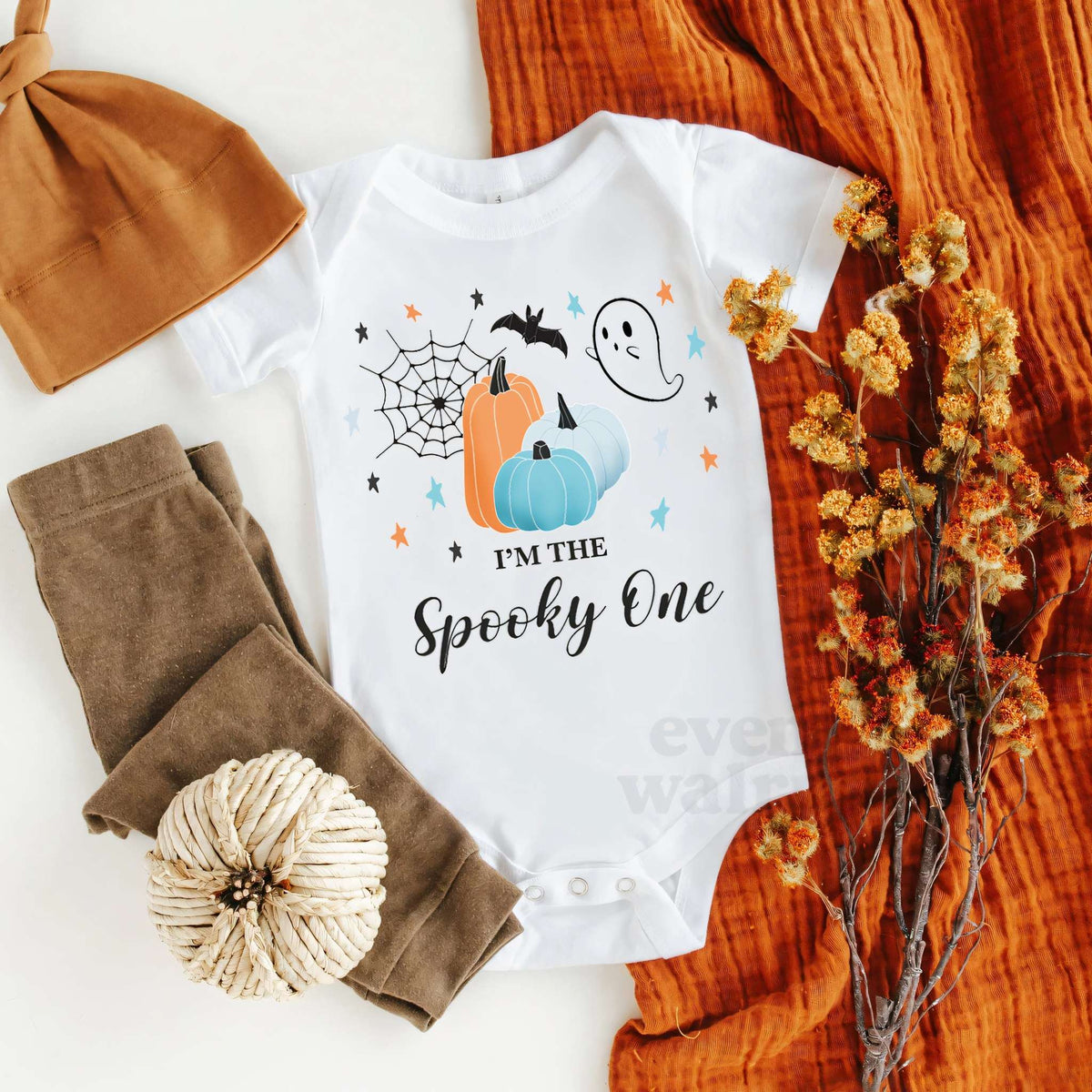 White I'm the Spooky One bodysuit laid flat with a knit hat, brown pants and fall leaves.