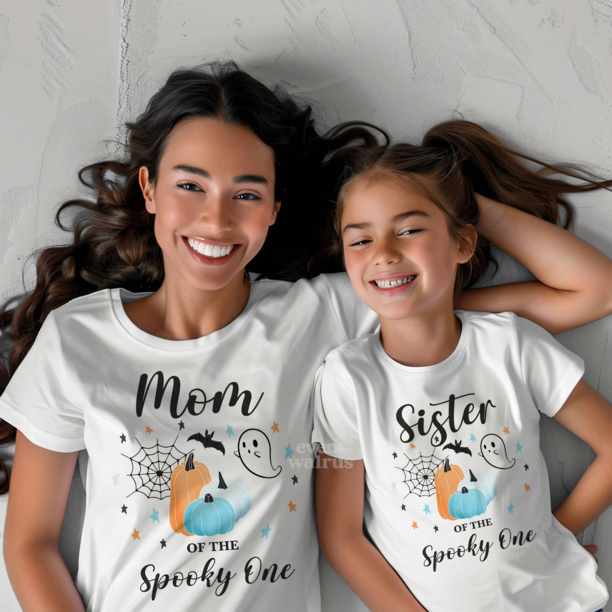 Little girl lying beside her mom, both in white Spooky One shirts.
