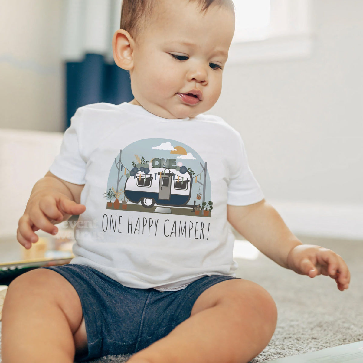 Baby boy sitting on a rug in a white One Happy Camper baby tee.