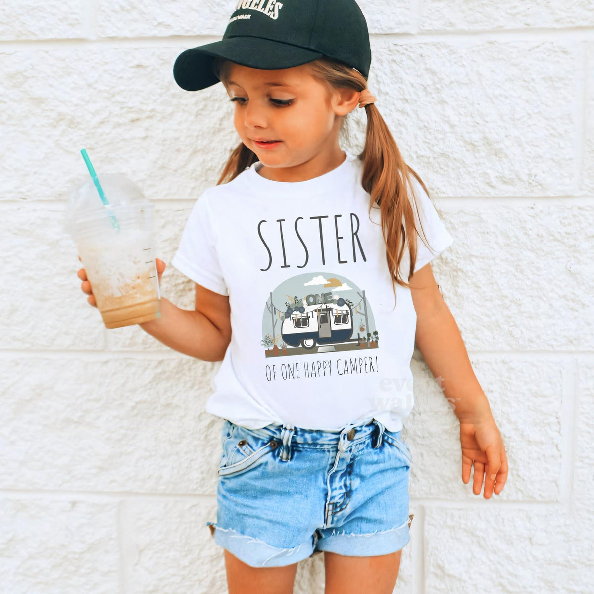 Girl in a ball cap holding a drink in a white Sister of One Happy Camper shirt.