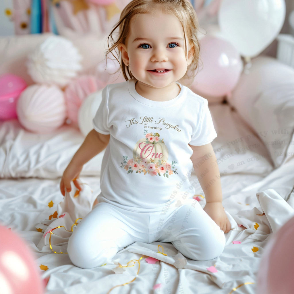 Smiling toddler girl among pink balloons in a white This Little Pumpkin Is Turning One tee.