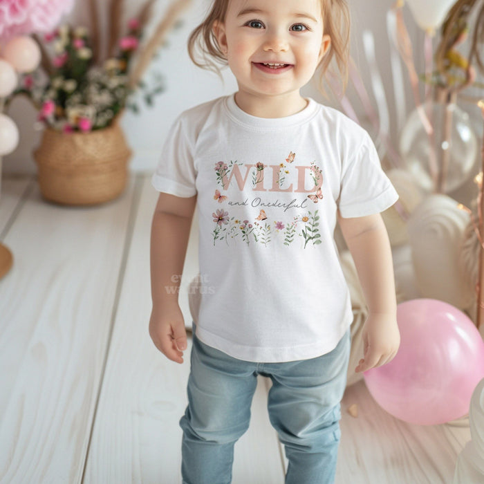 Smiling toddler girl at her party in a white Wild and Onederful tee with dusty pink wildflowers.