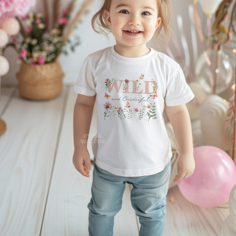 Smiling toddler girl at her party in a white Wild and Onederful tee with dusty pink wildflowers.