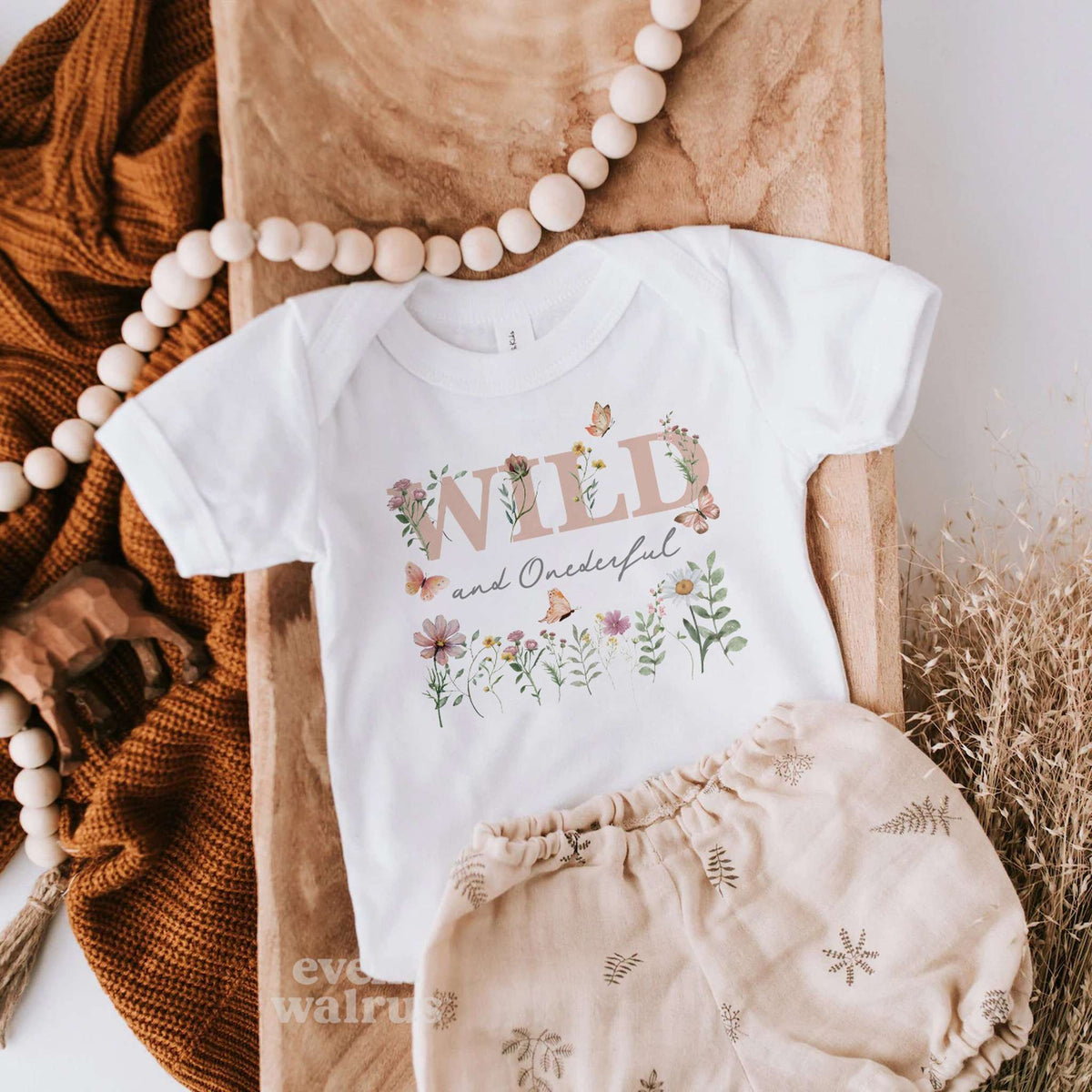 White Wild and Onederful baby bodysuit laid flat with linen bloomers and a wooden bead garland.