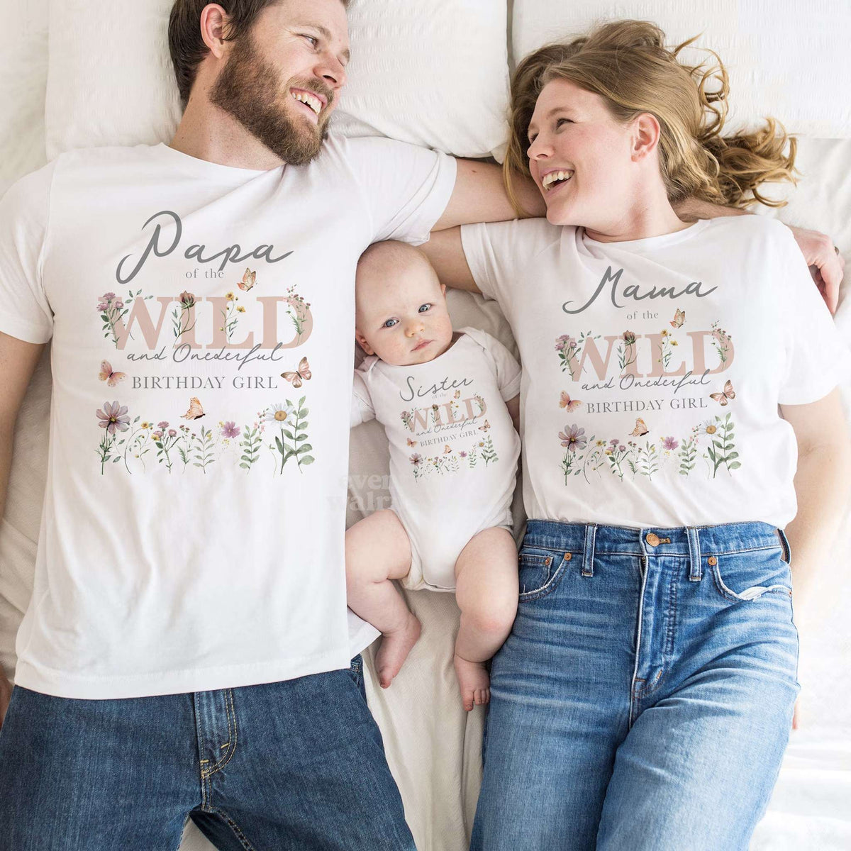 Parents lying beside their baby in a Sister of the Wild and Onederful bodysuit, all in white.