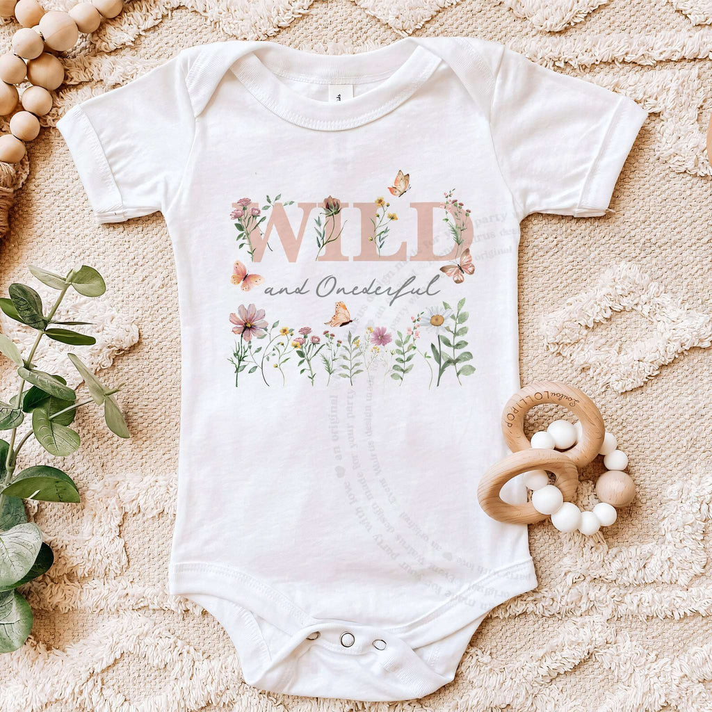 White Wild and Onederful first birthday bodysuit on a beige blanket with a wooden teether.