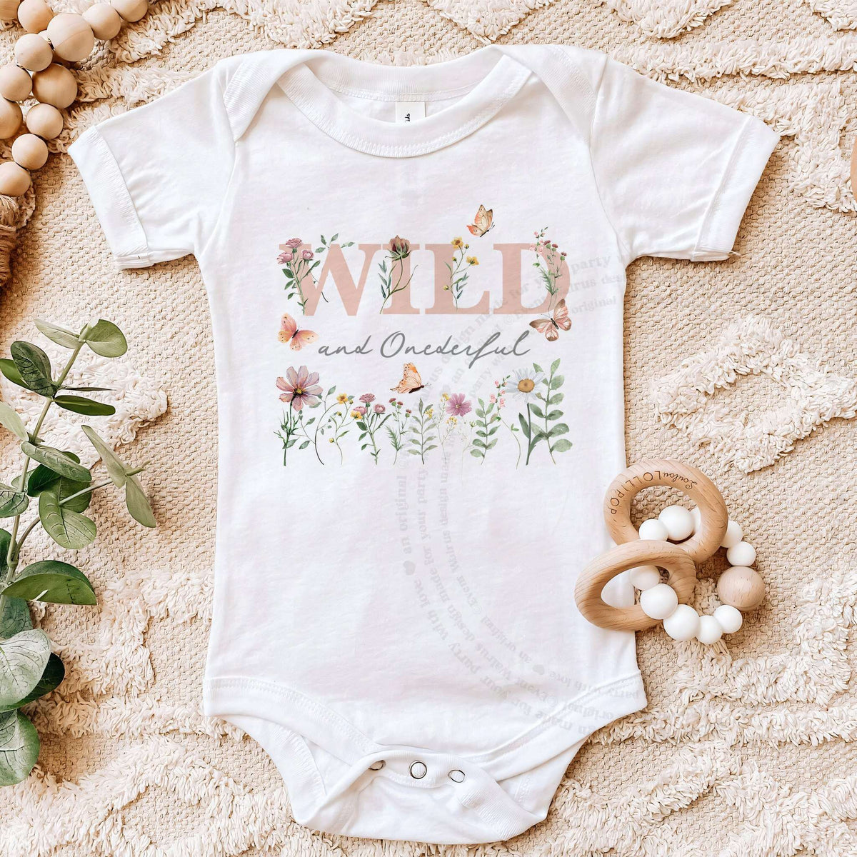 White Wild and Onederful first birthday bodysuit on a beige blanket with a wooden teether.