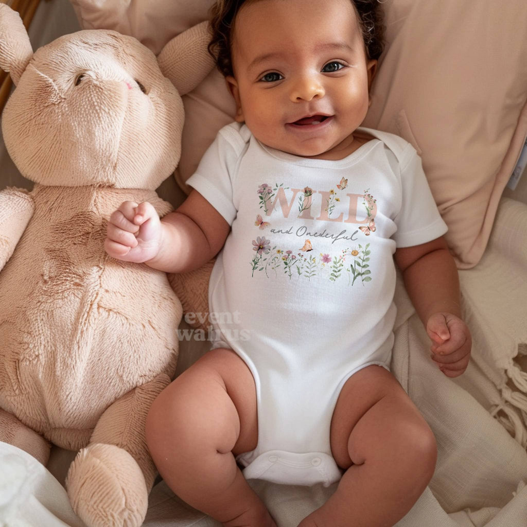 Smiling baby in a basket wearing a white Wild and Onederful bodysuit with pink wildflowers.