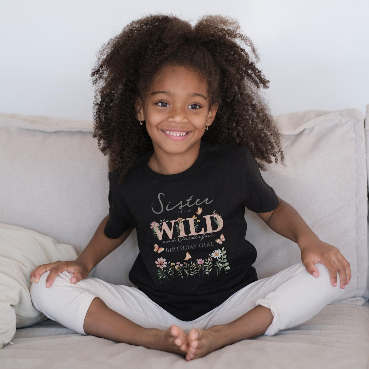 Smiling girl sitting on a couch in a black Sister of the Wild and Onederful tee.