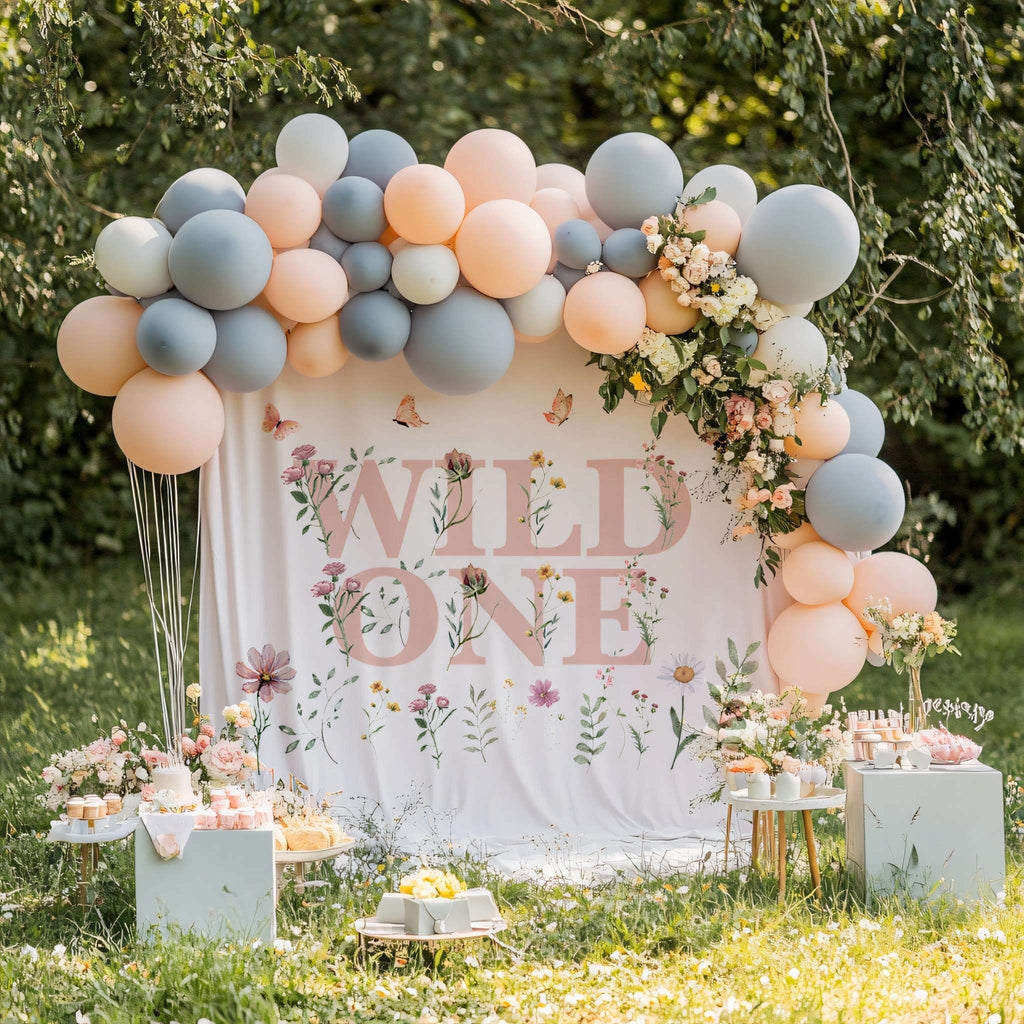 Wildflower Wild One backdrop behind a dessert table with a blue-gray and peach balloon arch.