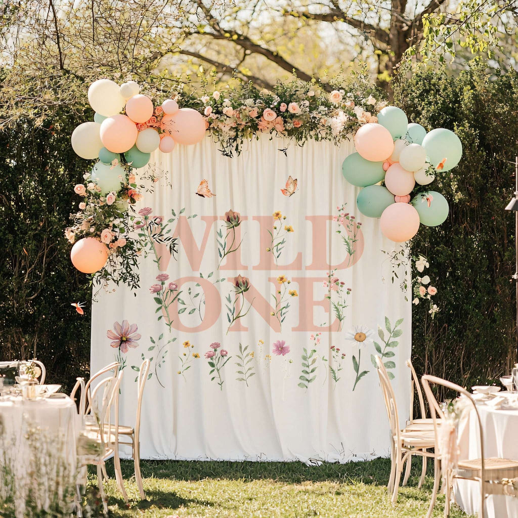 Wild One backdrop with pink wildflowers framed by balloon and flower garlands, with white party chairs.