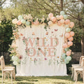 Pink wildflower Wild One backdrop in a garden with a flower garland and peach, sage and cream balloons.