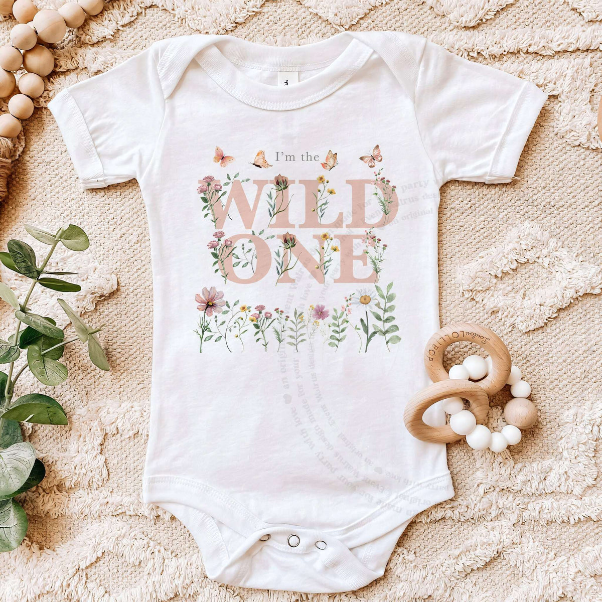 White I'm the Wild One first birthday bodysuit laid flat on a soft beige blanket with a wooden teether.