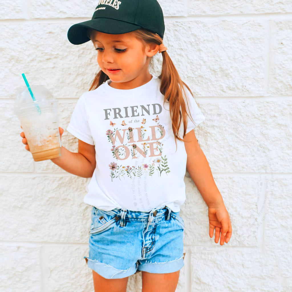 Girl holding a drink in a white Friend of the Wild One wildflower tee against a white brick wall.