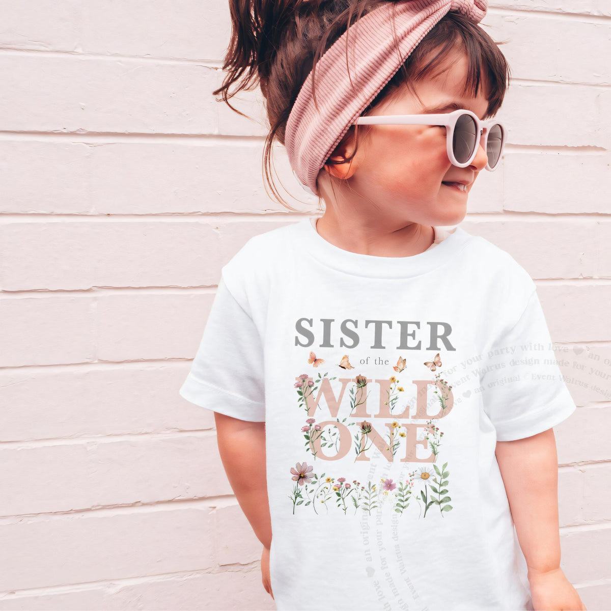 Girl in a white Sister of the Wild One tee and pink headband in front of a pink brick wall.