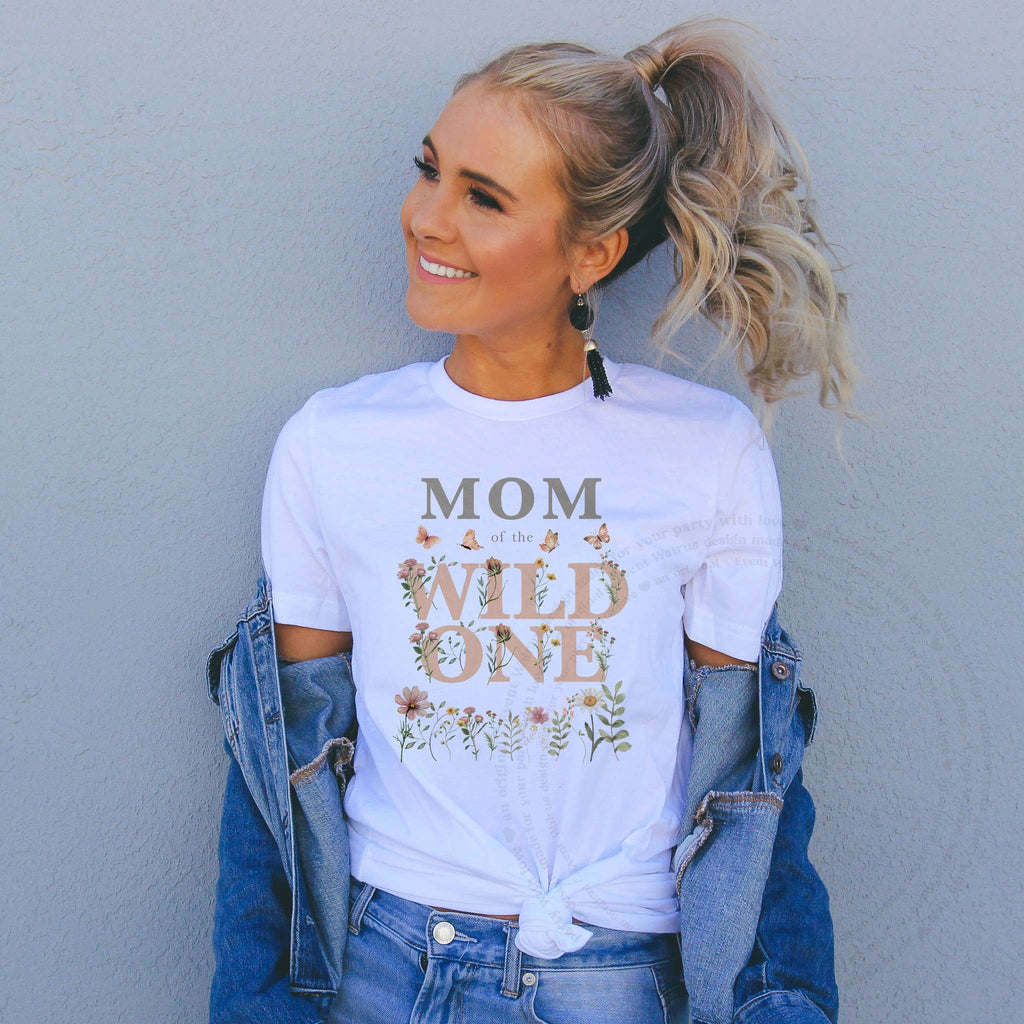 Smiling woman in a white Mom of the Wild One wildflower tee and denim jacket by a gray wall.