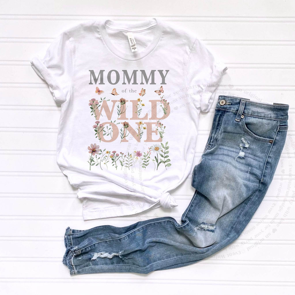 White Mommy of the Wild One adult tee laid flat beside jeans, with pink wildflower lettering.