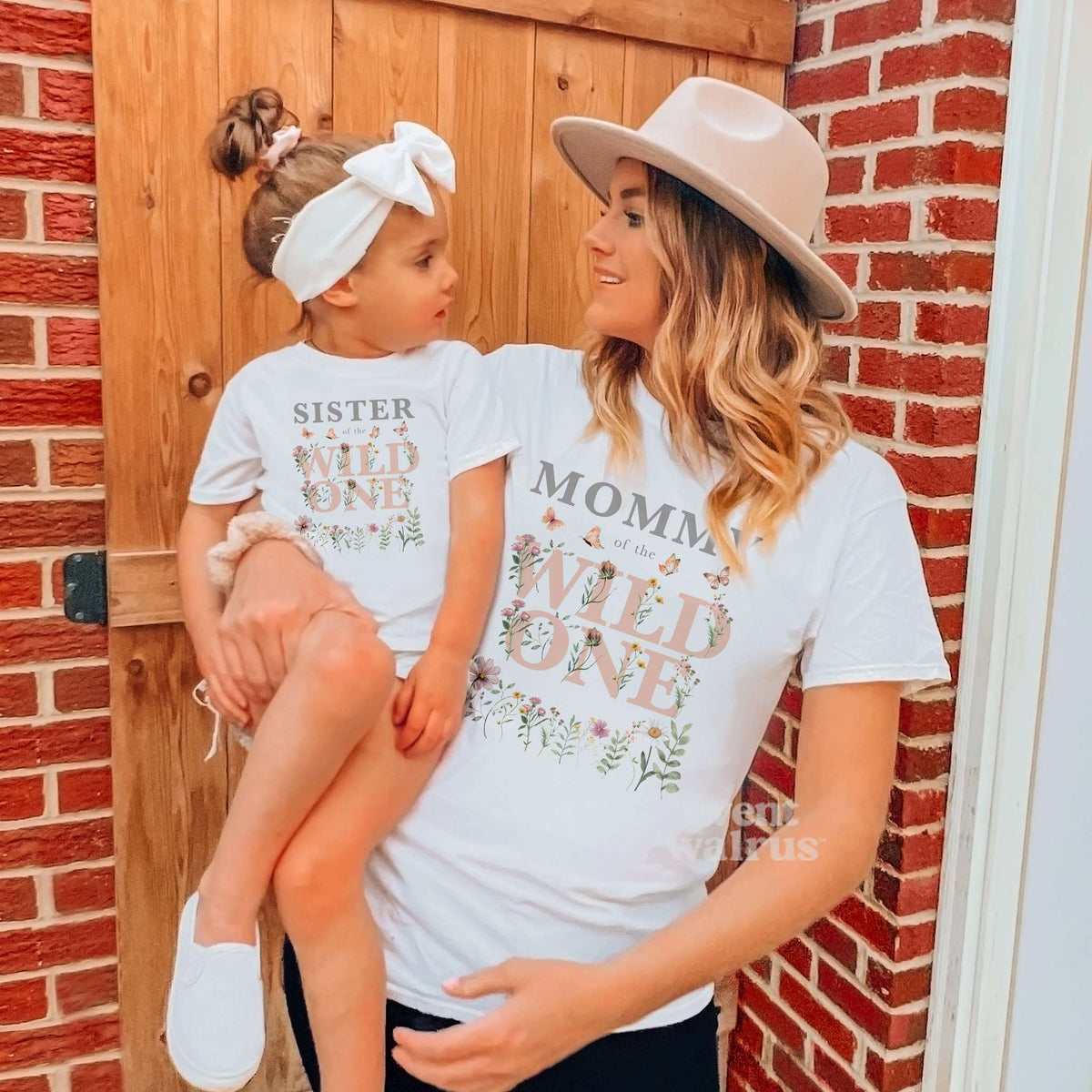 Mom holding her daughter, both in white Mommy and Sister of the Wild One wildflower shirts.