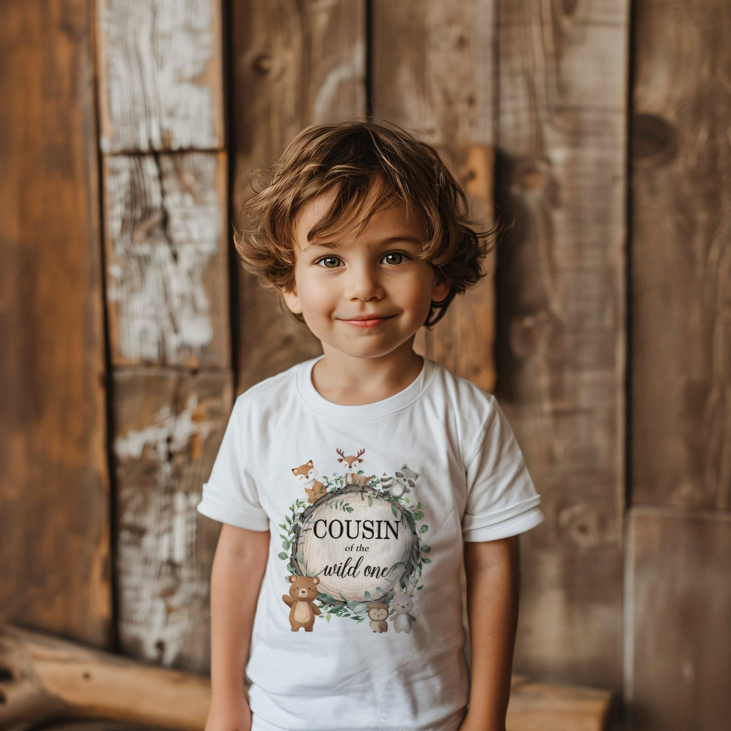 Toddler boy against a weathered wood wall in a white Cousin of the Wild One tee.