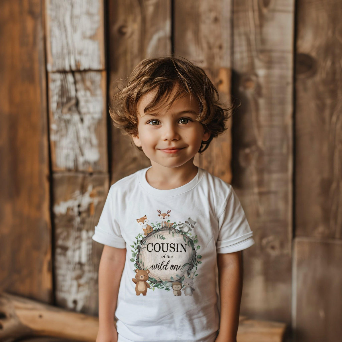 Toddler boy against a weathered wood wall in a white Cousin of the Wild One tee.