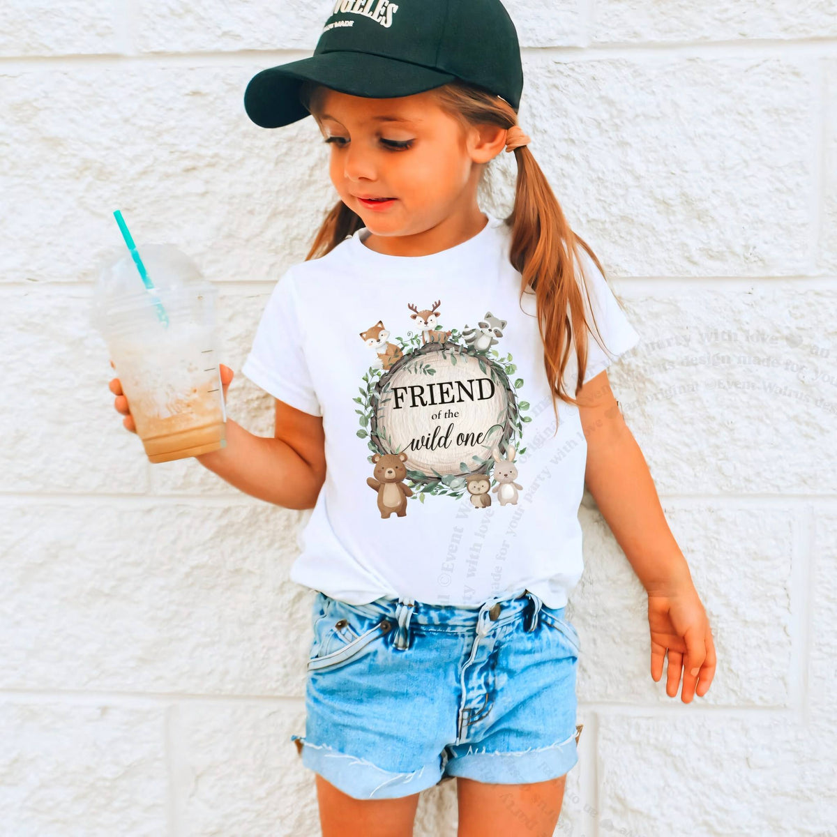 Girl in a ball cap holding a drink in a white Friend of the Wild One shirt.